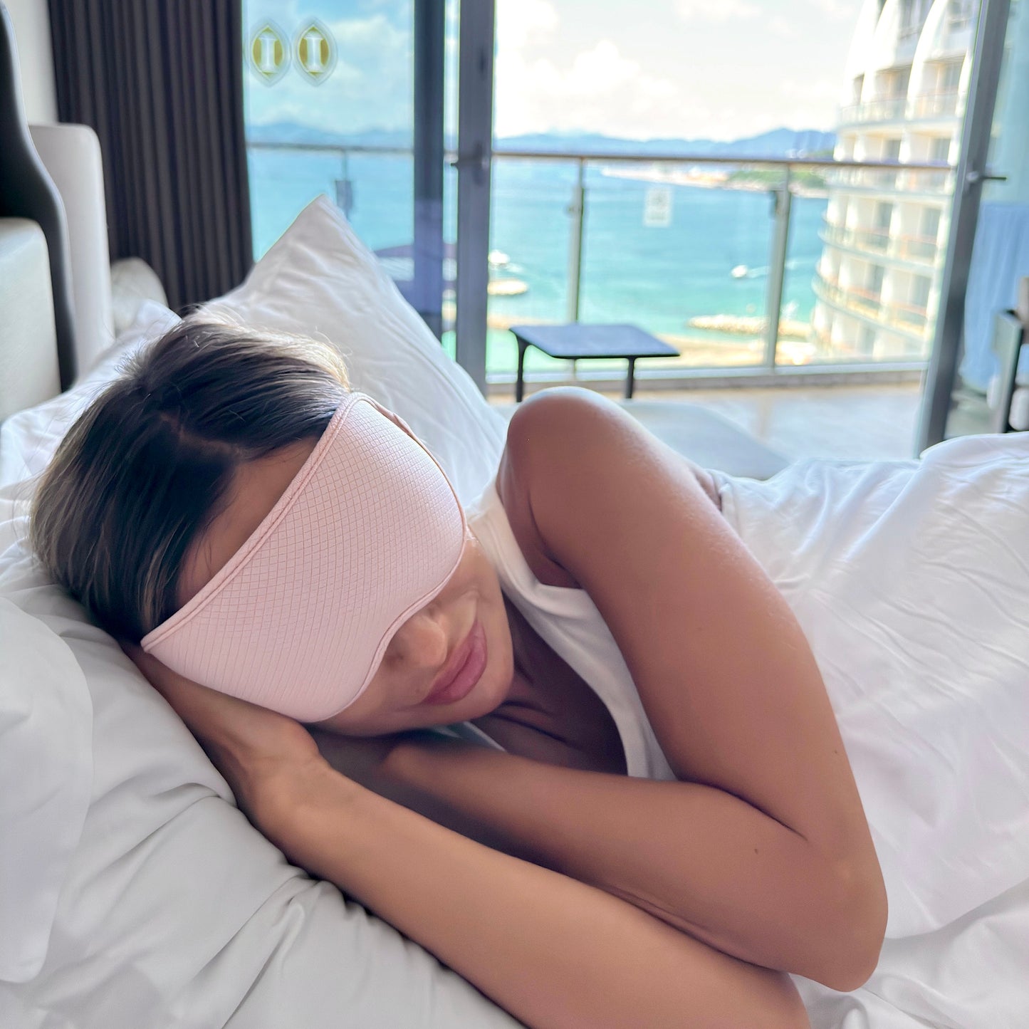 Woman relaxing in bed wearing LEVIQUE pink 3D sleep mask, comfortable for side sleepers, with ocean view background.