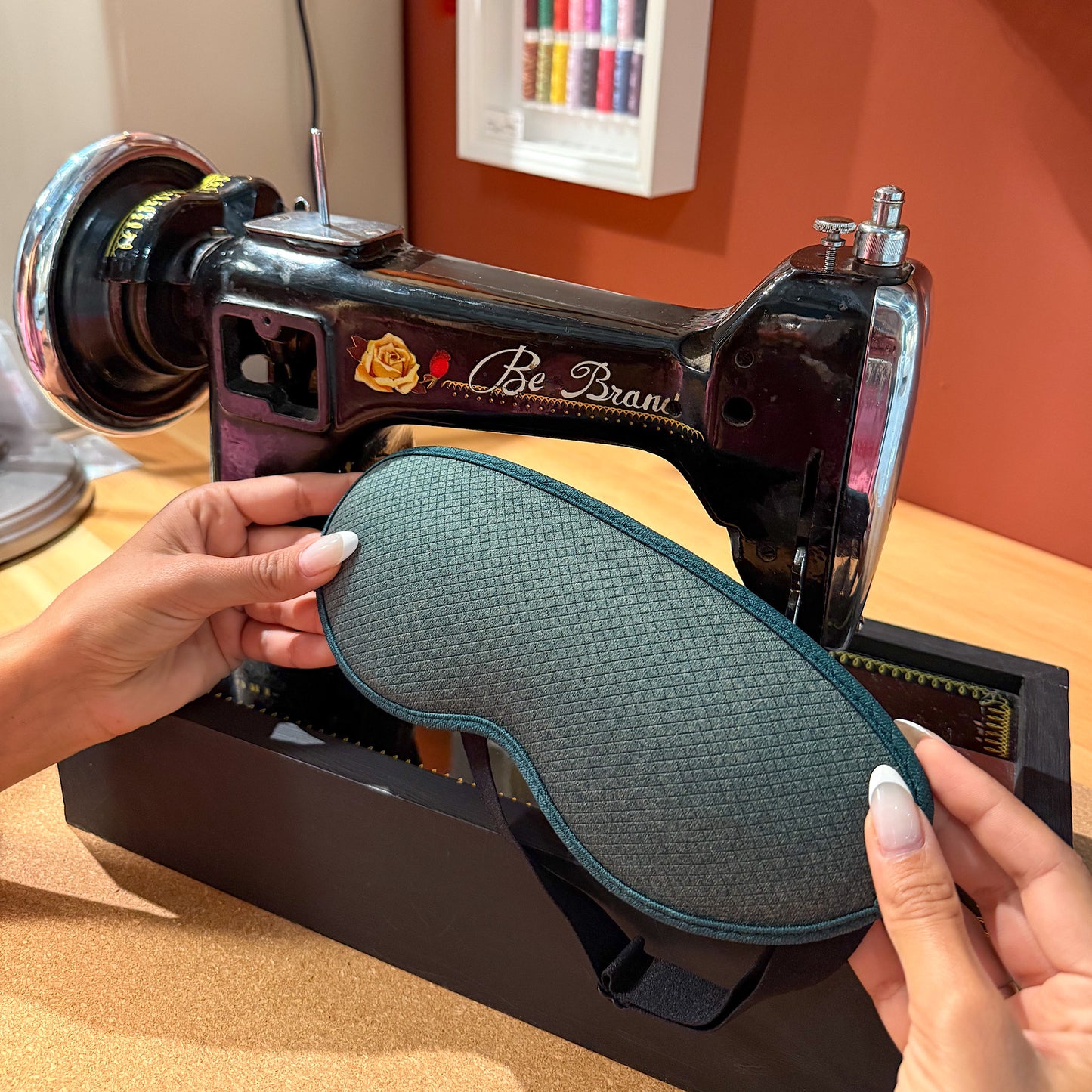 Close-up of LEVIQUE sleep mask being held in front of a vintage sewing machine, showcasing its high-quality material and design.