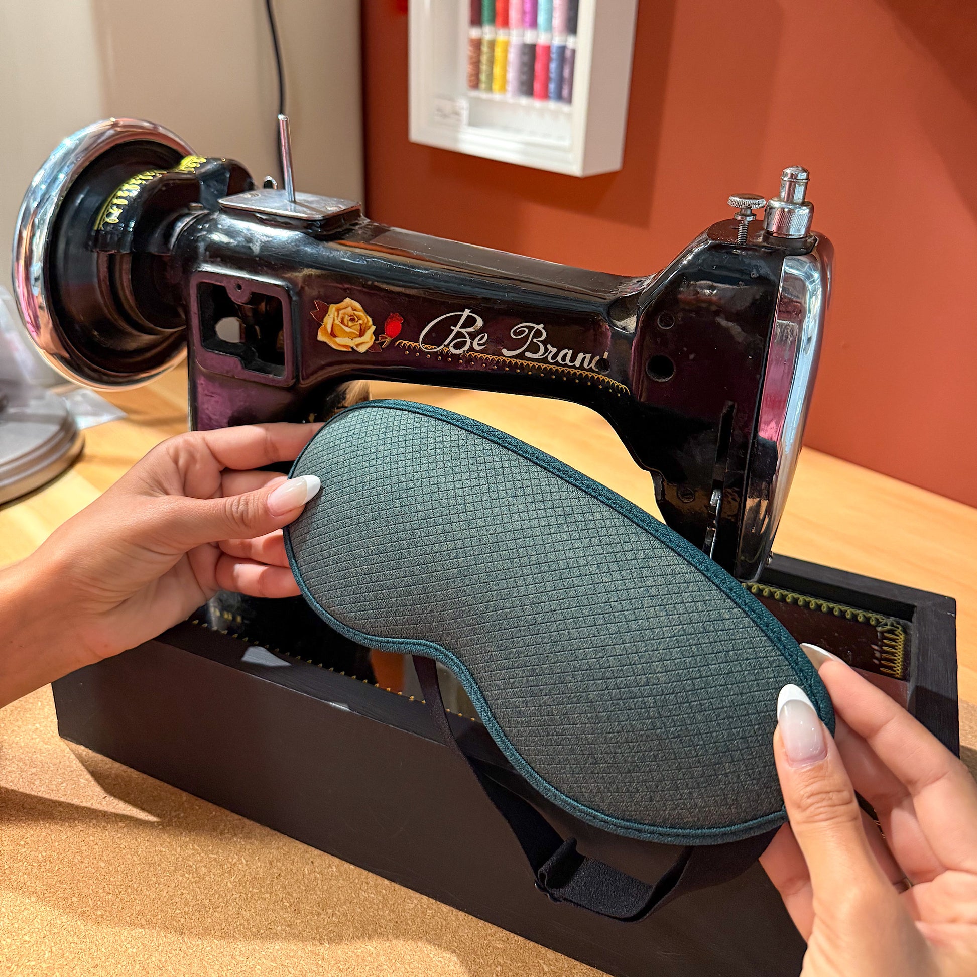 Close-up of LEVIQUE sleep mask being held in front of a vintage sewing machine, showcasing its high-quality material and design.